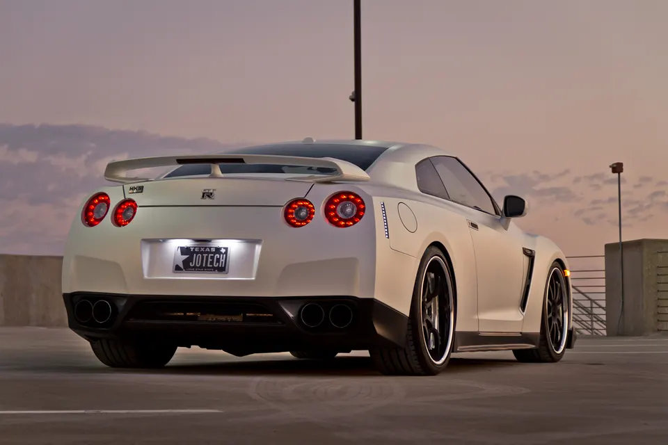 A white Nissan GT-R with aftermarket wheels and a custom Texas JOTECH license plate, parked at sunset.