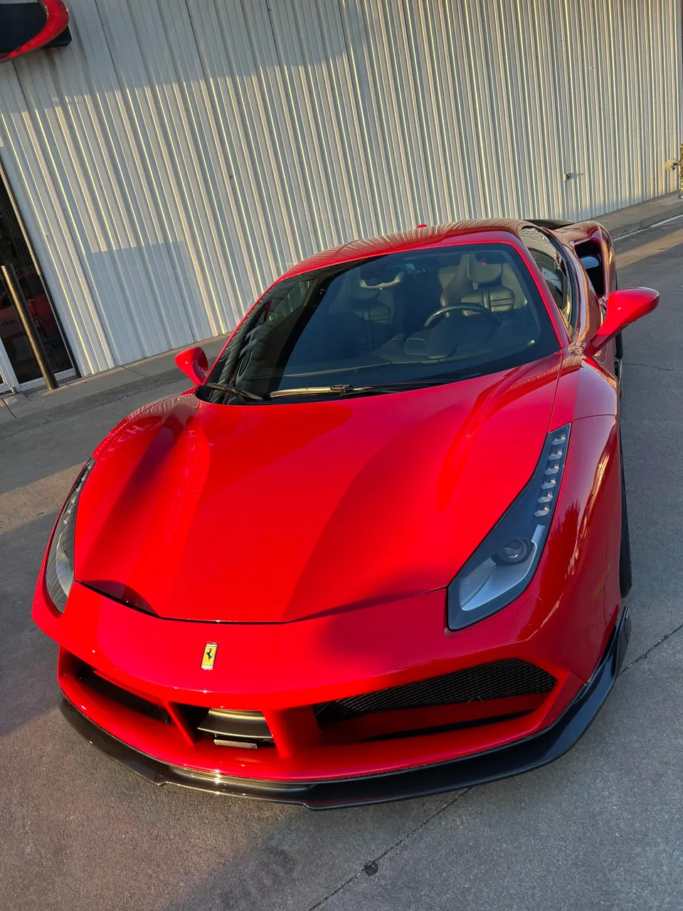 Red Ferrari 488 parked outside a building with a corrugated metal wall, showcasing its sleek design and carbon fiber front splitter.