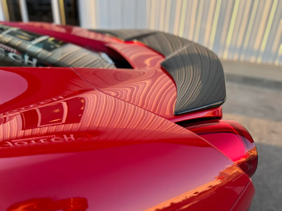 A close-up detail shot of a red sports car's rear, highlighting the carbon fiber aerodynamic wing and glossy paint finish.