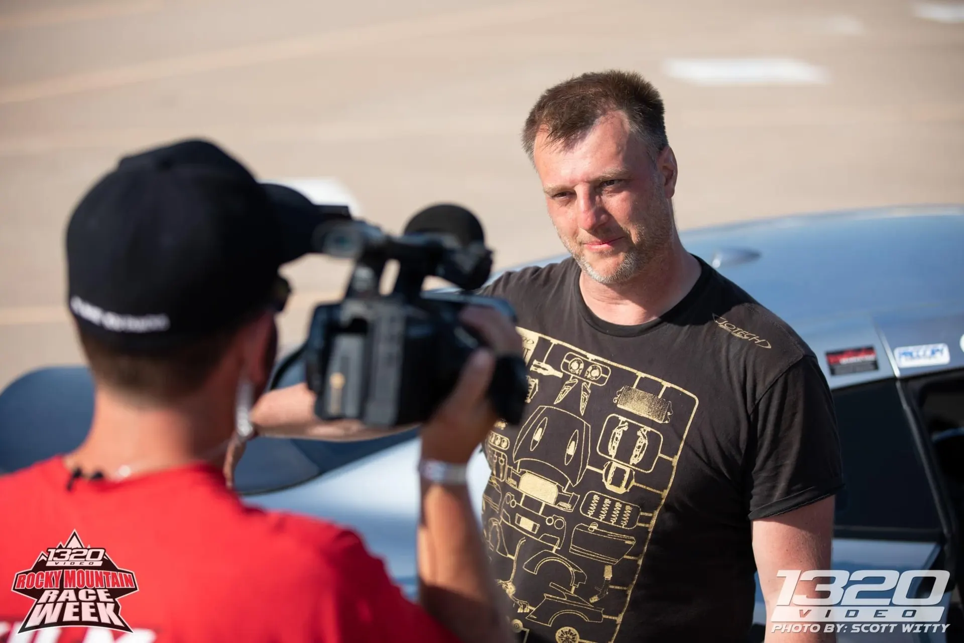 A videographer interviews a man at a Rocky Mountain Race Week event, with a sports car visible in the background.