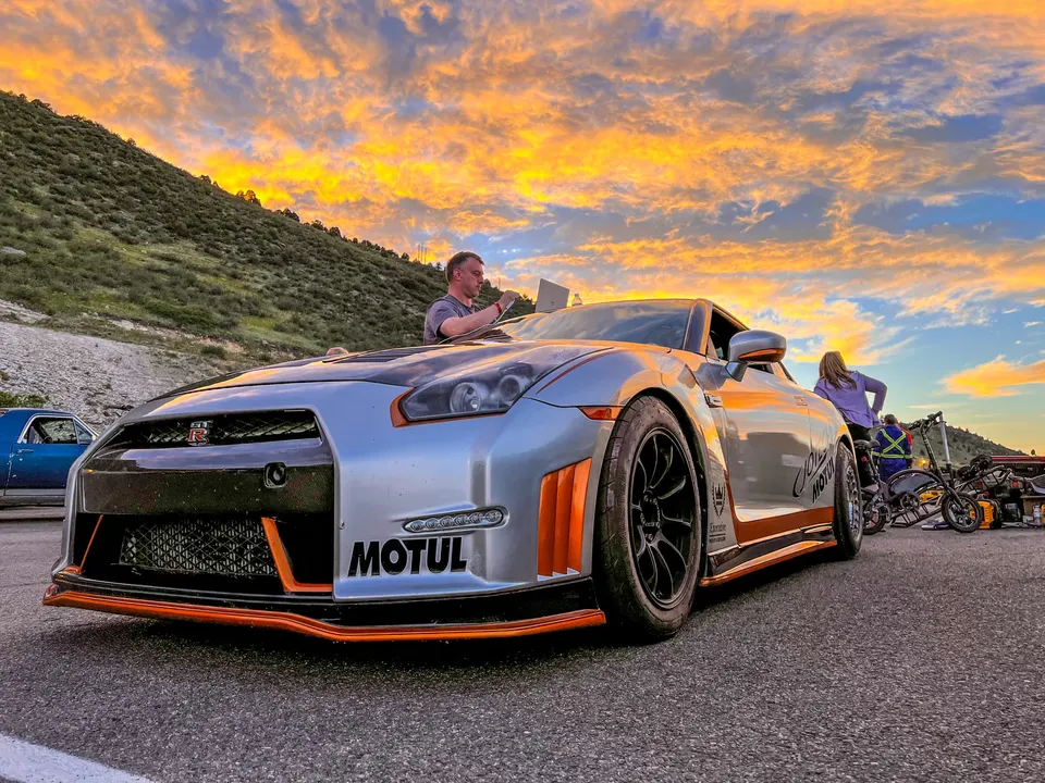 Silver Nissan GT-R with orange accents being tuned on a laptop at a track event during sunset.