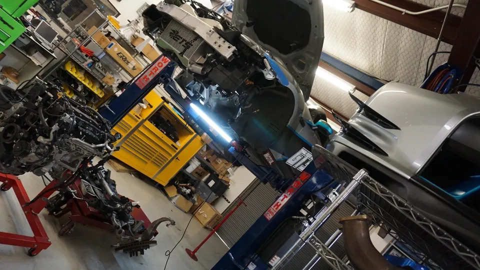 A Nissan GT-R on a lift in a performance tuning shop with its front end disassembled, an engine on a stand nearby.