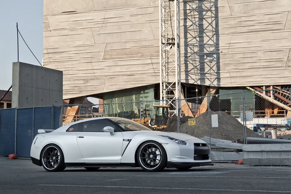 A white Nissan GT-R with aftermarket wheels parked in an urban setting with a modern building in the background.