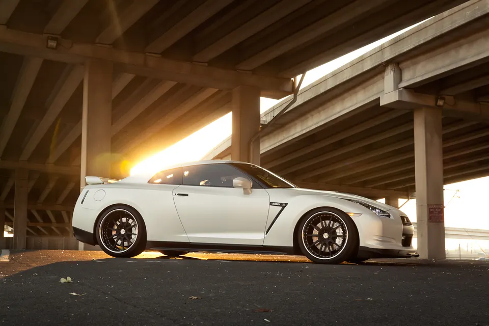 A white Nissan GT-R with custom black wheels parked under an overpass during golden hour, showcasing its sleek design.