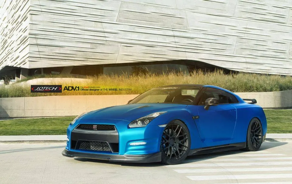 A blue Nissan GT-R with black ADV.1 wheels and a custom front splitter parked in front of a modern building.