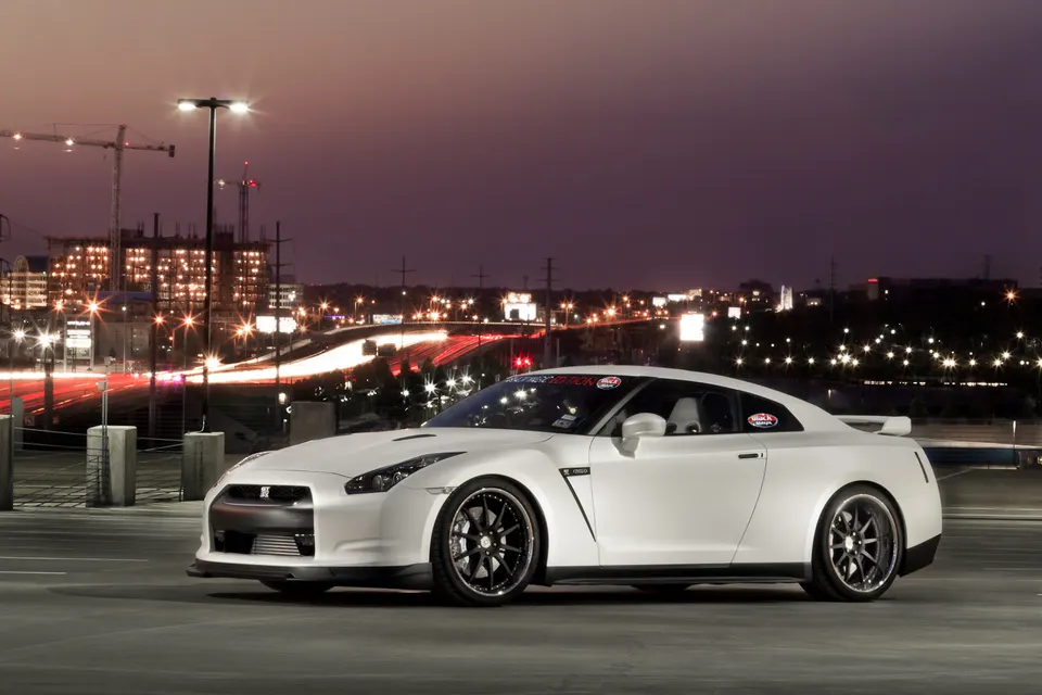 A white Nissan GT-R parked at night with city lights and light trails in the background, showcasing its sleek design.