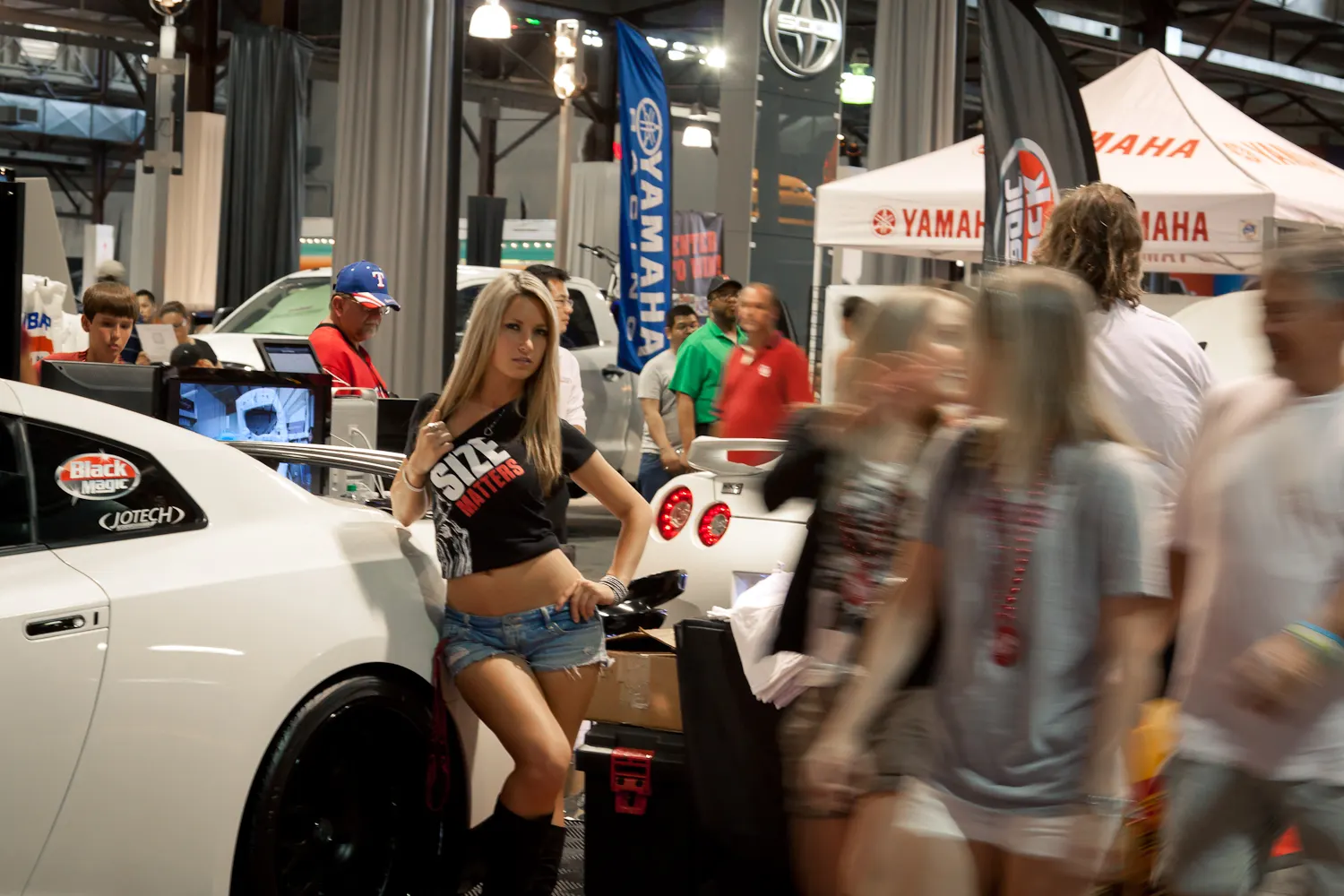 Woman posing next to a white Nissan GT-R at a car show event with blurred crowds and event banners in the background.