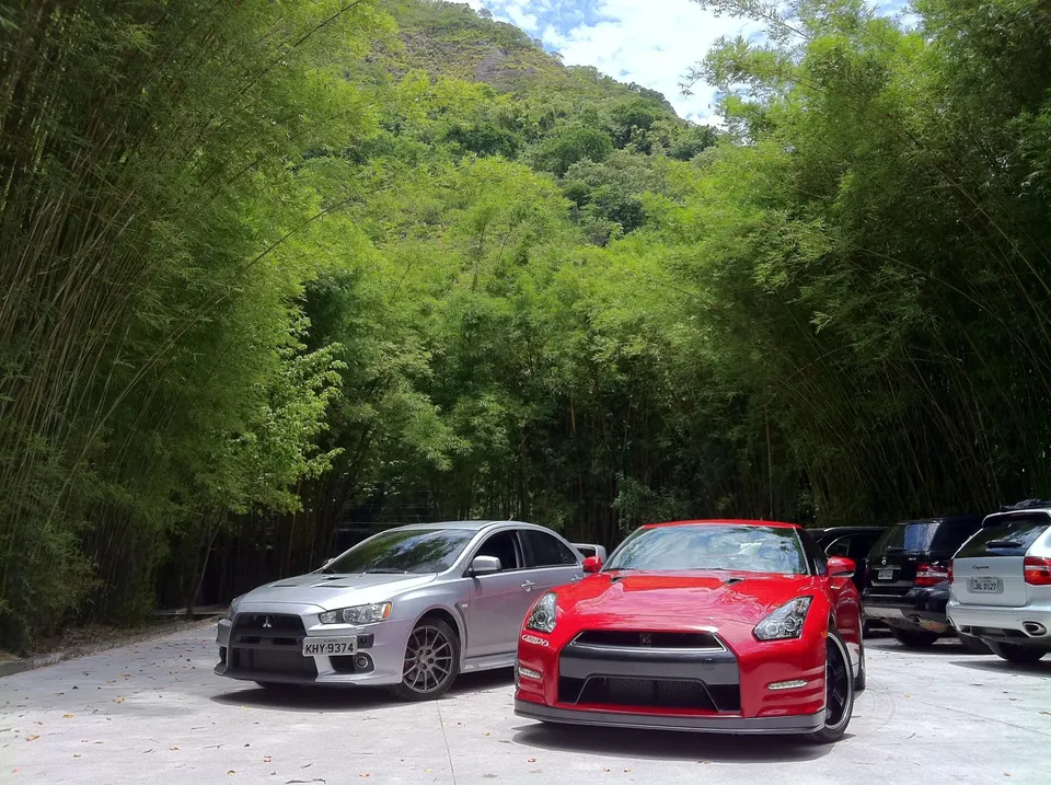 Red Nissan GT-R and silver Mitsubishi Lancer Evolution X parked outdoors among lush bamboo trees and a green mountain.