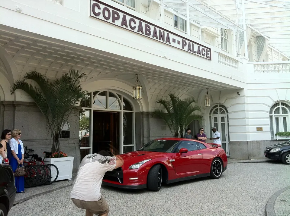 Red Nissan GT-R parked in front of the Copacabana Palace hotel with people around, one person taking a photo.