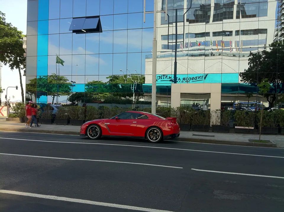 Red Nissan GT-R parked on a city street next to a modern building with reflective windows and a blue sky.