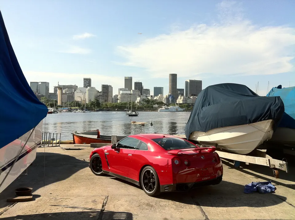 Red Nissan GT-R parked on a waterfront with a city skyline and boats in the background on a sunny day.