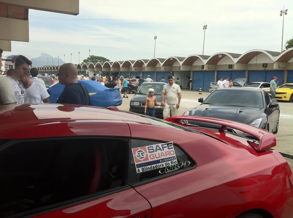 A red Nissan GT-R and a black Nissan GT-R parked at a car meet event at a racetrack, with people gathered around.