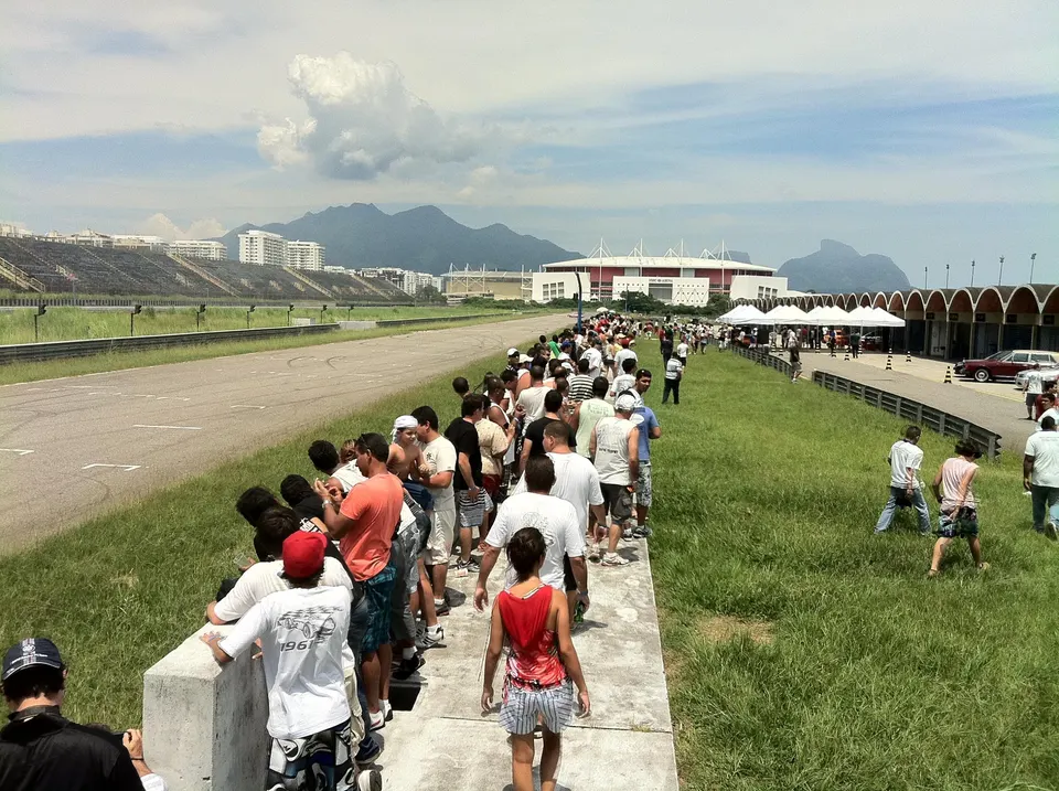 A large crowd of spectators lines a race track with grandstands and a stadium in the background under a cloudy sky.