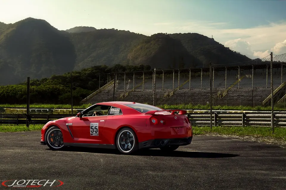 A red Nissan GT-R R35 race car with number 55 on a track, with grandstands and mountains in the background.