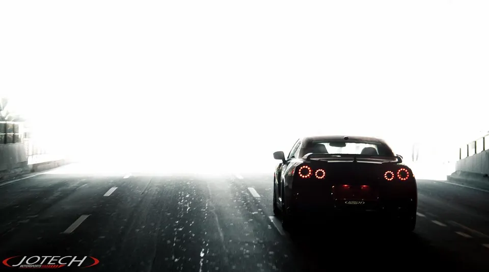 Rear view of a dark Nissan GT-R driving into a bright light at the end of a tunnel, showcasing its illuminated taillights.