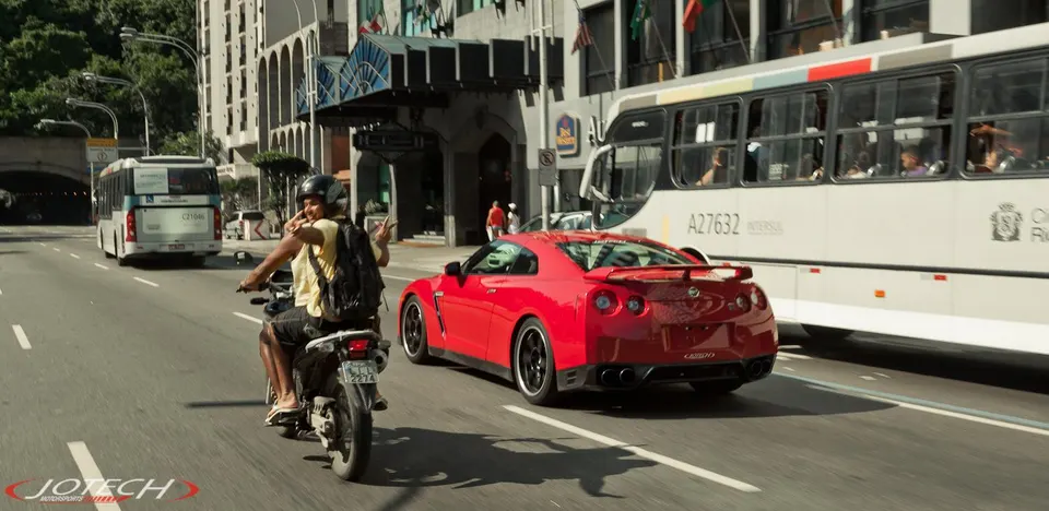 Red Nissan GT-R driving on a busy city street alongside a motorcycle and buses during the day.
