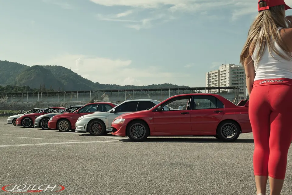 A lineup of Mitsubishi Evo and Subaru WRX cars parked on a track, with a woman in red leggings in the foreground.