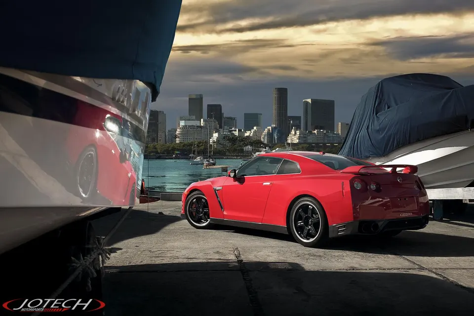 Red Nissan GT-R parked by a waterfront with a city skyline in the background, reflecting in a nearby boat.