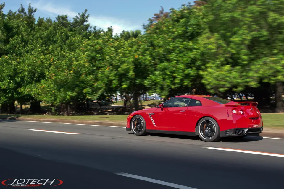 A red Nissan GT-R driving on a sunny road with trees in the background, showcasing its sleek design and aftermarket wheels.