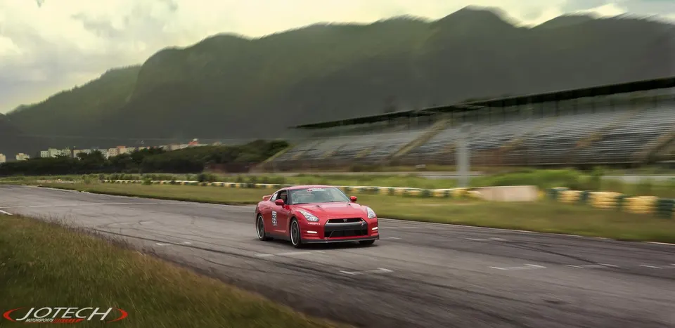 A red Nissan GT-R driving on a race track with mountains and grandstands in the background under a cloudy sky.