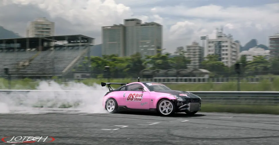 A pink Nissan 350Z drift car with a widebody kit and large rear wing performs a burnout on a race track.