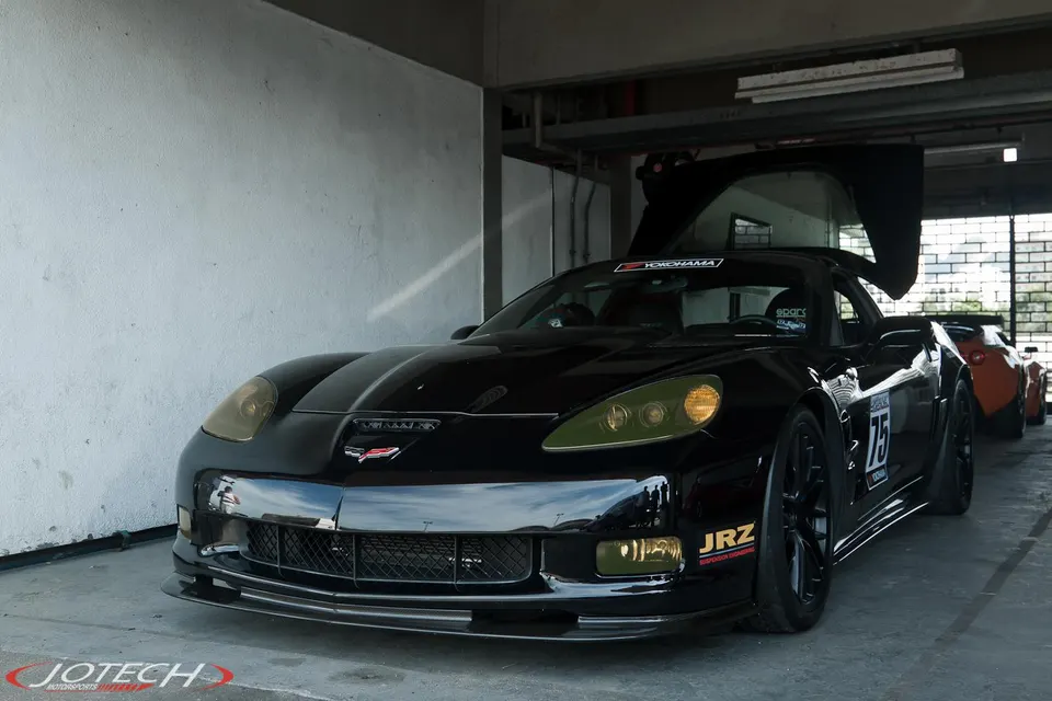 Black C6 Corvette race car with number 75 and JRZ suspension sticker parked in a garage or pit lane.