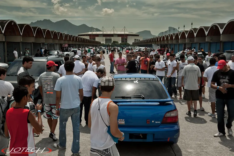 A large crowd of people gathered around a blue Subaru WRX and other cars at an outdoor car meet with a stadium in the background.