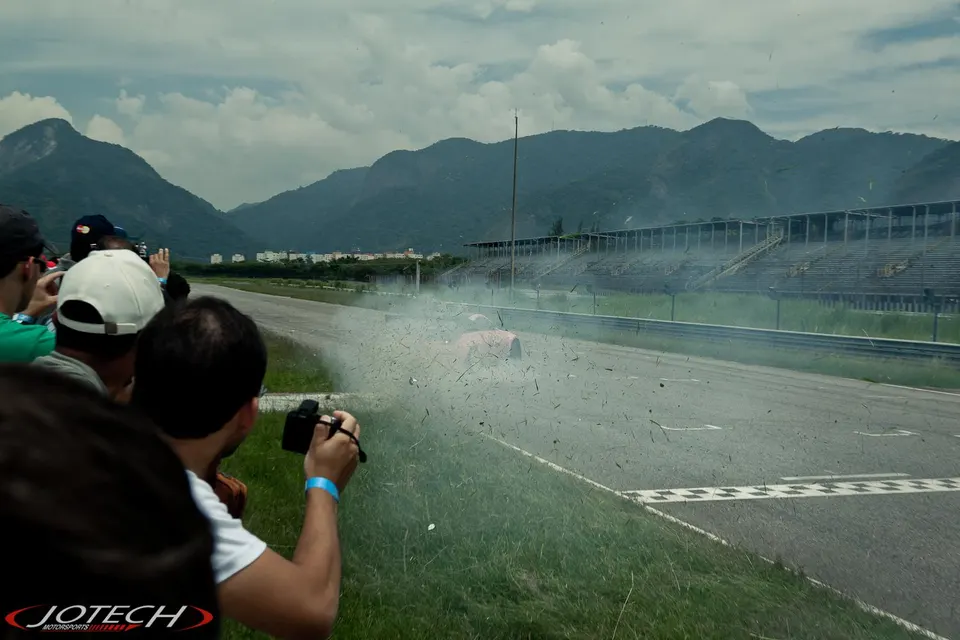 Spectators watch a car on a race track engulfed in smoke and debris after an incident, with mountains in the background.