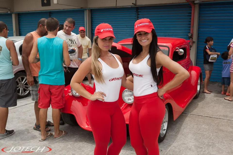 Two women in red hats and white tops with "Oktane" logo pose in front of a red classic car at an outdoor event.