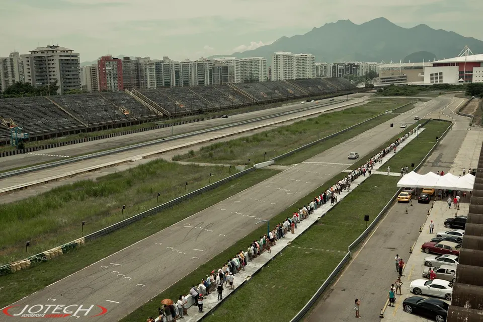 Aerial view of a race track with grandstands, a long line of spectators, and cars on the track and parked.