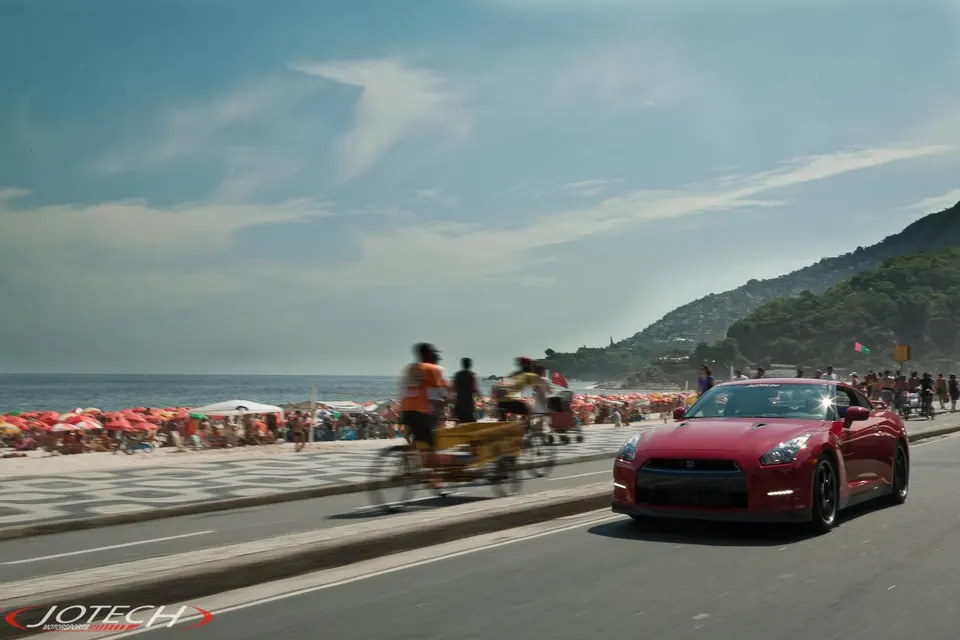 Red Nissan GT-R driving on a coastal road next to a busy beach with umbrellas and people under a blue sky.