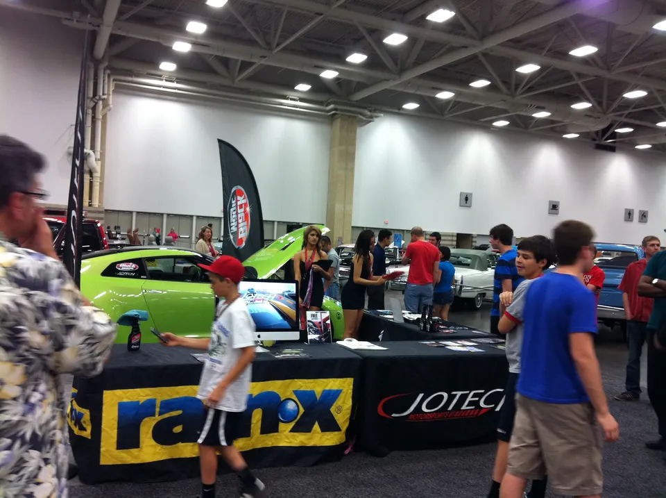 Attendees gather around a lime green car with its hood open at an indoor car show, with Jotech and Ranx displays.