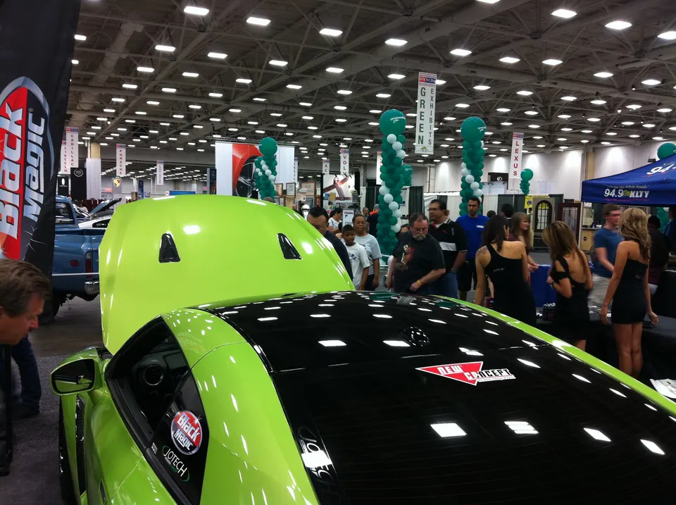 A bright green sports car with its hood open on display at a car show, surrounded by attendees and vendor booths.