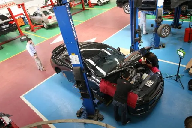 Mechanics working on a black car's engine bay in a busy auto repair shop with multiple car lifts and a customer observing.