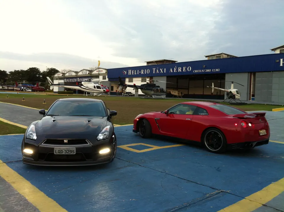 Two Nissan GT-Rs, one black and one red, parked on a tarmac in front of a heliport hangar with helicopters.