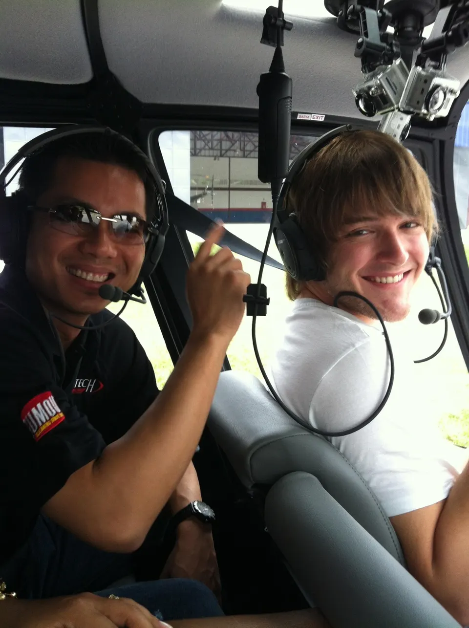 Two men wearing headsets and smiling inside a helicopter cockpit with cameras mounted on the ceiling.