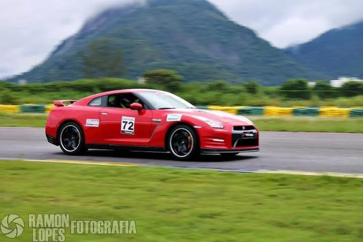 A red Nissan GT-R race car with number 72 on its side speeds around a turn on a race track with mountains in the background.