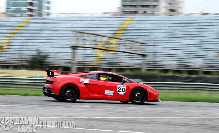 A red Lamborghini Gallardo race car with number 20 on the side speeds around a track with grandstands in the background.