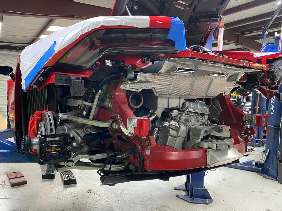 Red Corvette C8 on a lift in a performance shop, showing exposed rear suspension, brakes, and mid-engine components.
