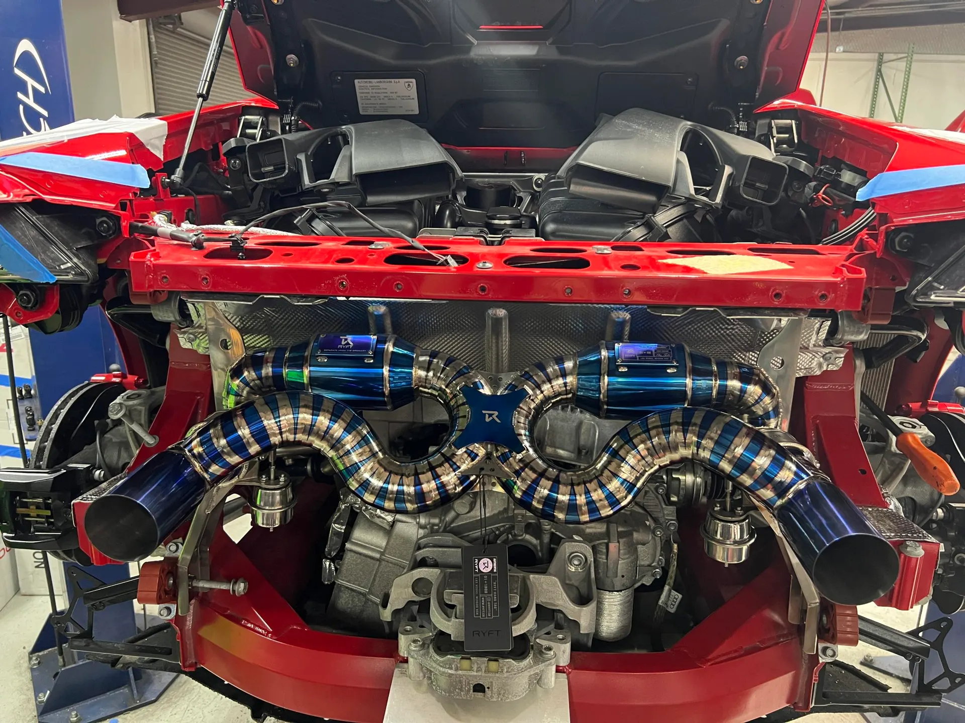 A red Lamborghini with its rear open, showcasing a custom titanium exhaust system installed in a performance shop.
