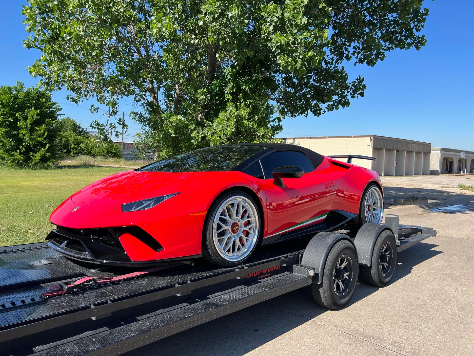 Red Lamborghini Huracan convertible with aftermarket wheels and a rear wing, loaded on a car trailer outdoors.