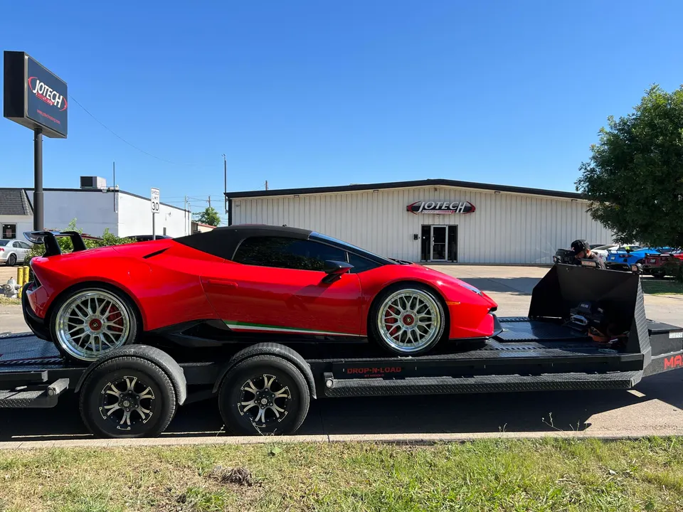 Red Lamborghini Huracan convertible on a car hauler trailer in front of Jotech Motorsports shop building.