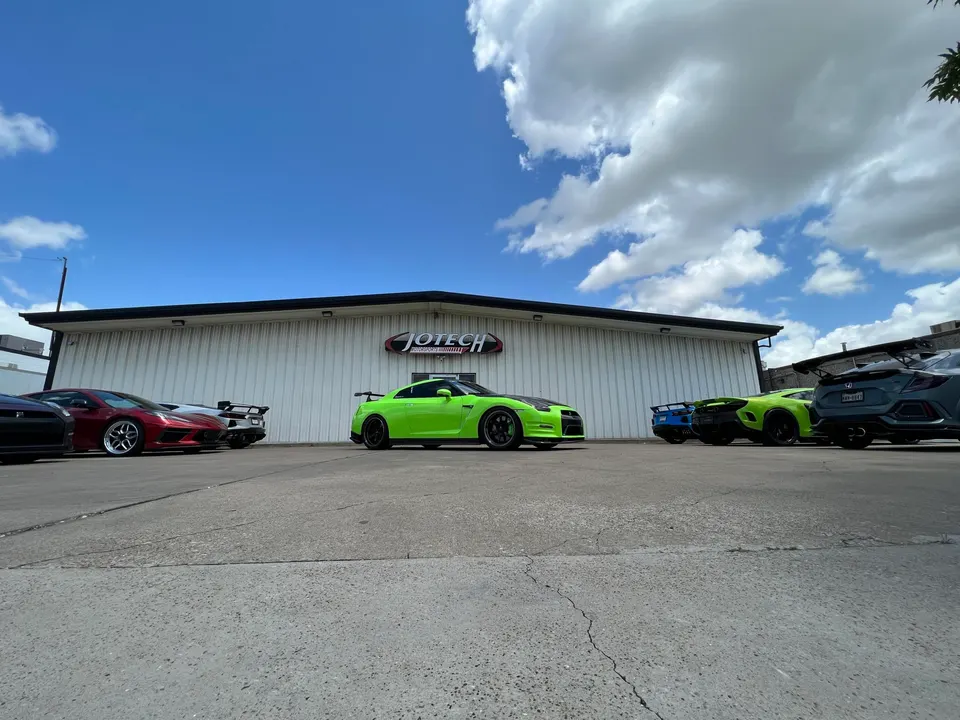 Multiple performance cars including a green Nissan GT-R and McLaren parked outside Jotech Motorsports tuning shop.
