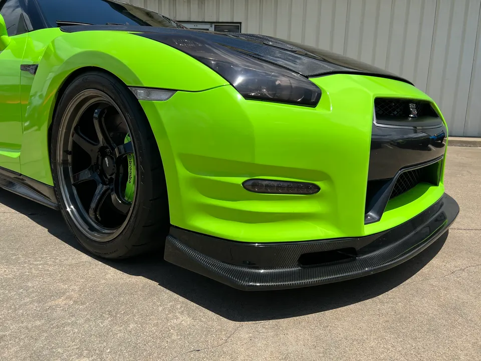Close-up of a lime green Nissan GT-R featuring a carbon fiber front splitter, aftermarket black wheels, and Brembo brakes.
