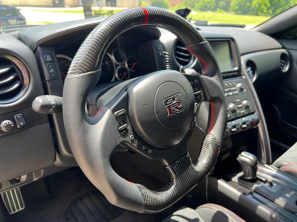 Close-up of a custom carbon fiber steering wheel with red stitching and a red stripe in a Nissan GT-R interior.