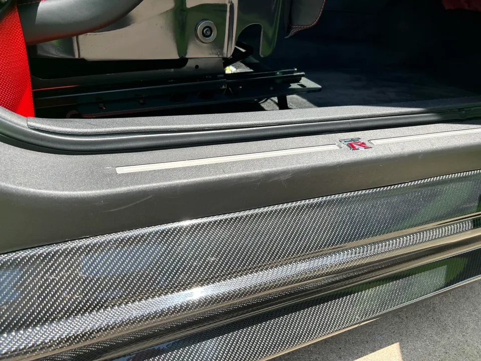 Close-up of a Nissan GT-R interior showing the carbon fiber door sill with GT-R logo and a red seatbelt.