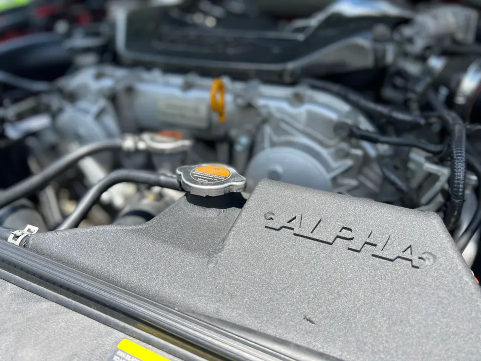 Close-up of a car engine bay featuring an Alpha Performance aftermarket intake system and radiator cap.