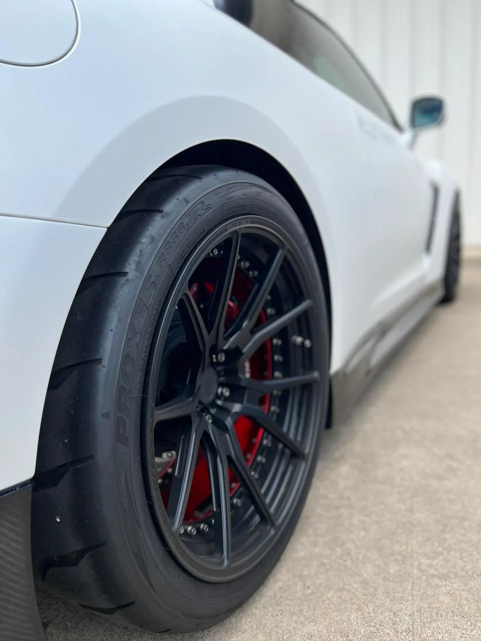 A close-up of the rear wheel of a white Nissan GT-R, featuring black multi-spoke wheels, red brake calipers, and Toyo Proxes R888R tires.