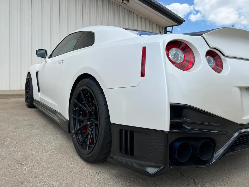 White Nissan GT-R with aftermarket black wheels, carbon fiber side skirts, and a rear diffuser with blue exhaust tips.