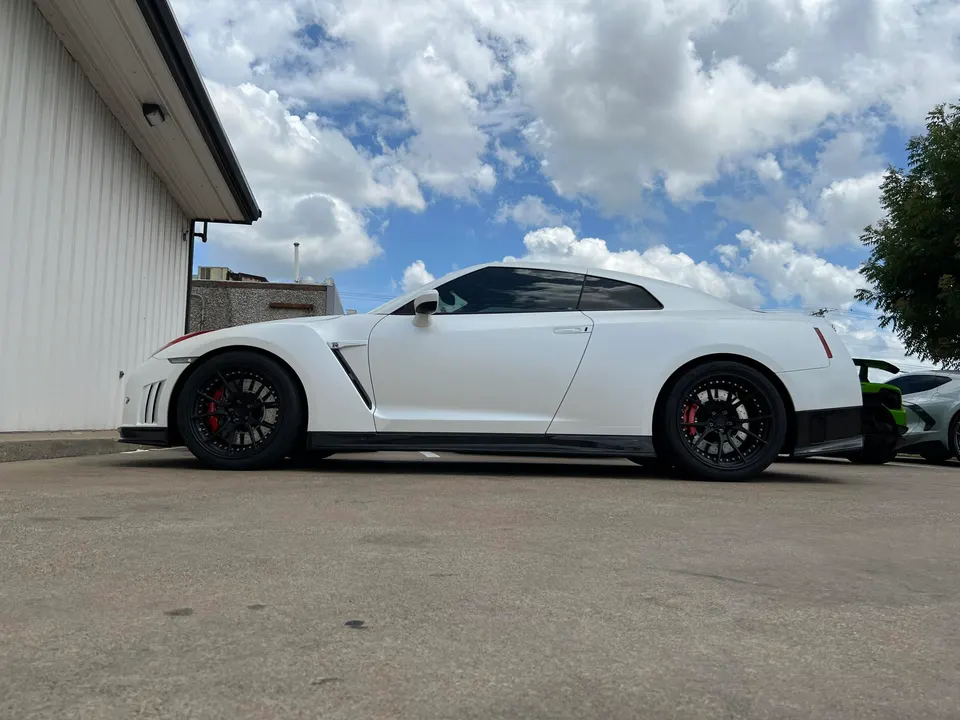 A white Nissan GT-R with aftermarket black wheels and red brake calipers parked outside a building under a cloudy sky.